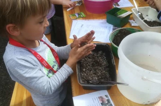 Ein Kind formt Erde mit den H&auml;nden an einem Tisch voller Materialien. Es tr&auml;gt ein rotes Band um den Hals. Verschiedene Beh&auml;lter und Anleitungen sind zu sehen., &copy; PlochingenInfo