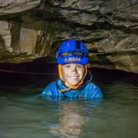 Eine Person mit Helm und Stirnlampe steht lächelnd in einer wassergefüllten Höhle. Die Umgebung ist von Felsen umgeben., © SwabianTravel