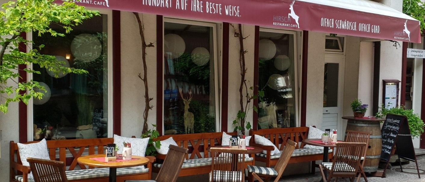 Gemütlicher Außenbereich des Restaurant Café Hirsch mit Holzbänken, Tischen und Pflanzen. Ein rotes Vordach mit Schriftzug ist sichtbar., © Restaurant Café Hirsch Gemütlicher Außenbereich des Restaurant Café Hirsch mit Holzbänken, Tischen und Pflanzen. Ein rotes Vordach mit Schriftzug ist sichtbar., © Restaurant Café Hirsch