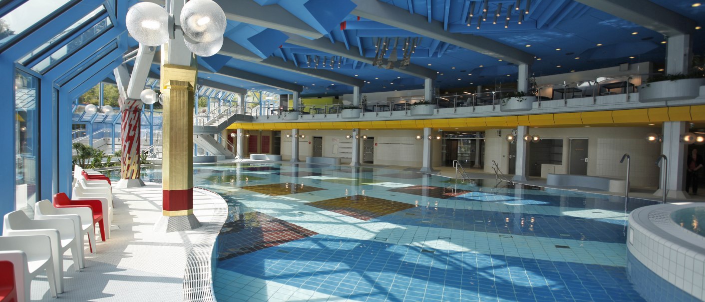 Modernes Schwimmbad mit blauer Decke, bunten Fliesen und weißen sowie roten Stühlen entlang des Beckens., © Stuttgarter Bäder