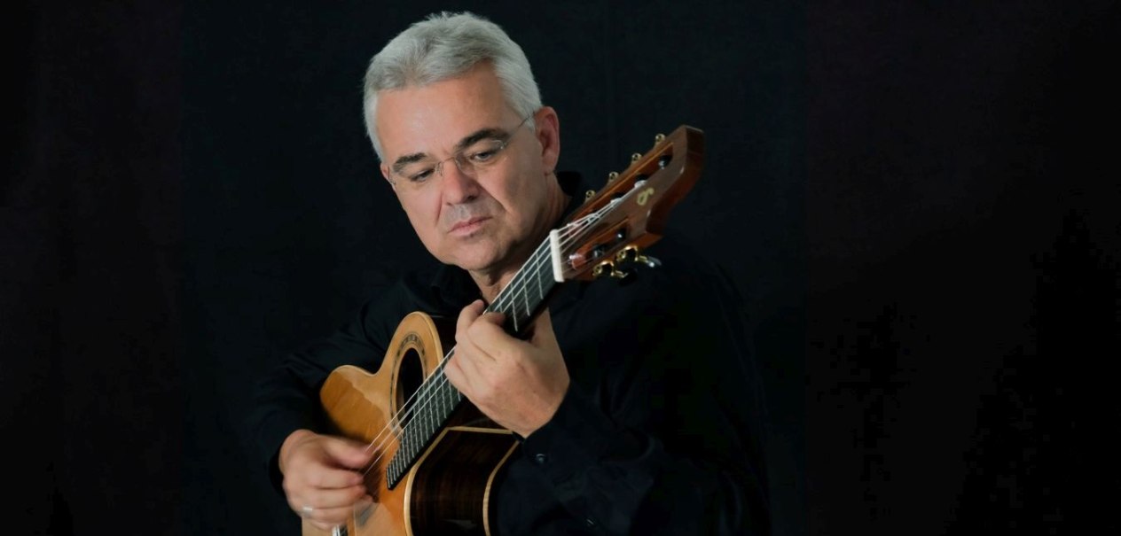 Mann mit grauen, kurzgeschnittenen Haaren und Brille vor schwarzem Hintergrund, auf einer Gitarre spielend, © Volker Luft