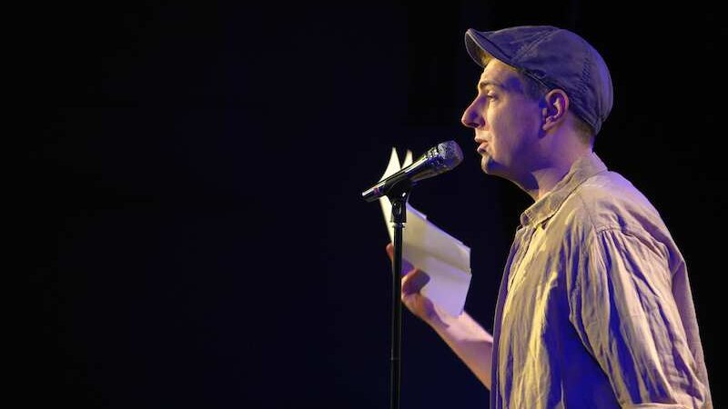 Ein Mann mit M&uuml;tze spricht in ein Mikrofon und h&auml;lt ein Blatt Papier. Er steht auf einer B&uuml;hne, vermutlich bei einem Poetry Slam., &copy; Kulturhaus Schwanen
