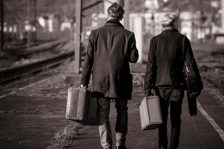 Zwei Männer in Mänteln tragen Koffer und gehen auf einem verlassenen Bahnsteig entlang. Die Szene ist in Schwarz-Weiß gehalten., © FMS Photography