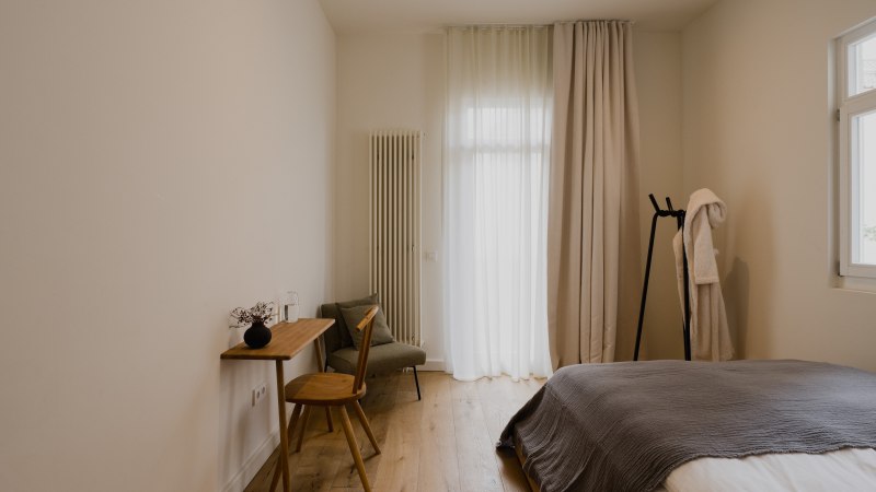 Minimalistisch eingerichtetes Schlafzimmer mit Holzboden, Bett, kleinem Schreibtisch und Stuhl. Vorhänge und ein Bademantel auf einem Ständer ergänzen das Bild., © Peter Oliver Wolff / DAS EDITH Minimalistisch eingerichtetes Schlafzimmer mit Holzboden, Bett, kleinem Schreibtisch und Stuhl. Vorhänge und ein Bademantel auf einem Ständer ergänzen das Bild., © Peter Oliver Wolff / DAS EDITH