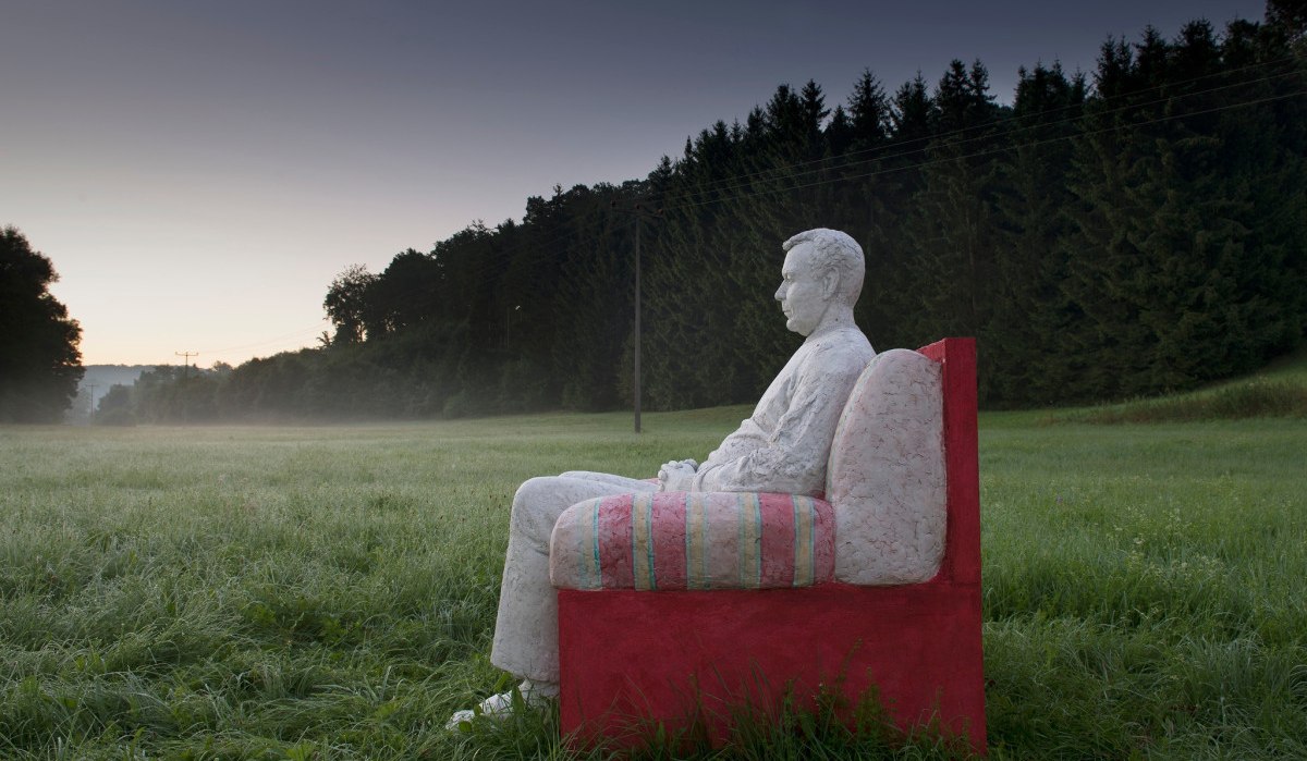 Eine weiße Skulptur eines Mannes sitzt auf einem roten Sofa in einer grünen Wiese, umgeben von Bäumen im Hintergrund., © Natur.Nah. Schönbuch & Heckengäu Eine weiße Skulptur eines Mannes sitzt auf einem roten Sofa in einer grünen Wiese, umgeben von Bäumen im Hintergrund., © Natur.Nah. Schönbuch & Heckengäu