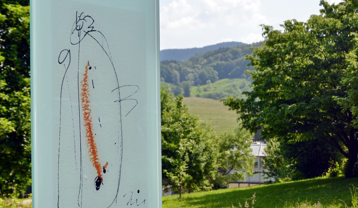Ein Kunstwerk auf Glas zeigt abstrakte Linien und Farben. Im Hintergrund ist eine grüne Landschaft mit Bäumen und Hügeln zu sehen., © Foto: Cornelia Steinbach Ein Kunstwerk auf Glas zeigt abstrakte Linien und Farben. Im Hintergrund ist eine grüne Landschaft mit Bäumen und Hügeln zu sehen., © Foto: Cornelia Steinbach
