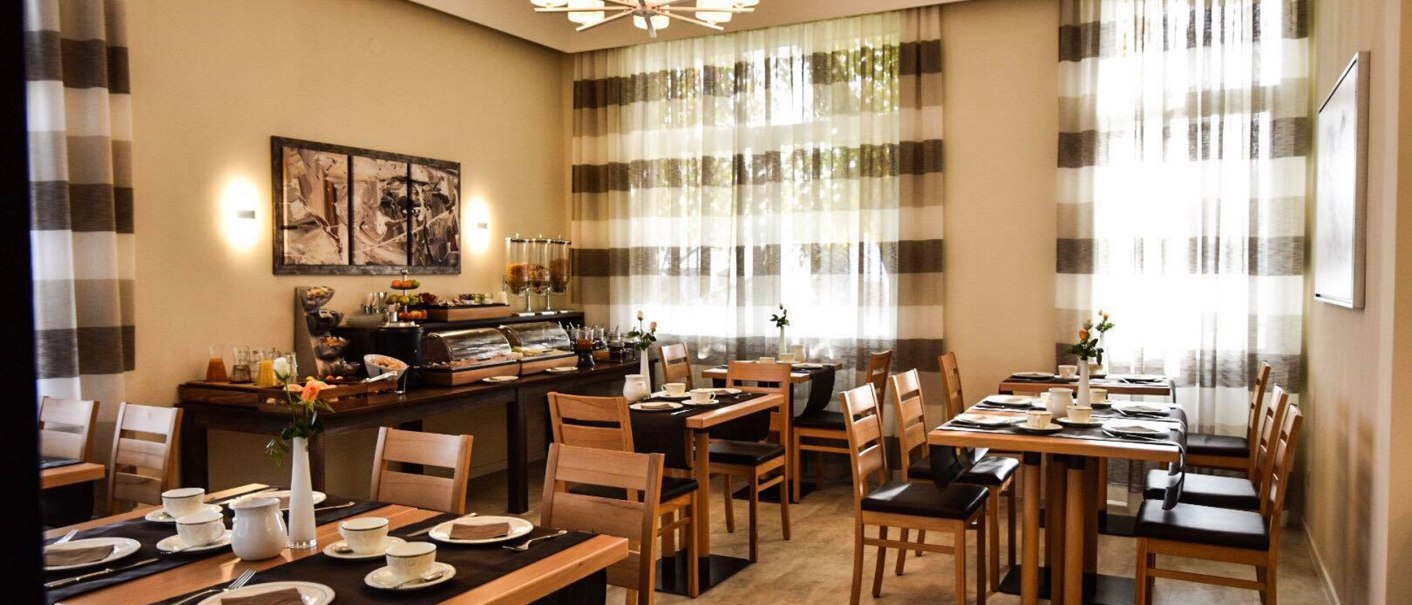 Ein eleganter Frühstücksraum mit Holzmöbeln, gedeckten Tischen und einem Buffet. Große Fenster mit gestreiften Vorhängen lassen Licht herein., © TOMAS Ein eleganter Frühstücksraum mit Holzmöbeln, gedeckten Tischen und einem Buffet. Große Fenster mit gestreiften Vorhängen lassen Licht herein., © TOMAS