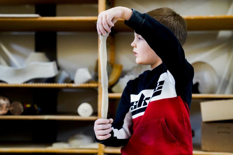 Ein Junge in einem rot-schwarzen Pullover formt Ton in einem Workshop. Im Hintergrund sind Regale mit Materialien zu sehen., © Kunstschule Labyrinth