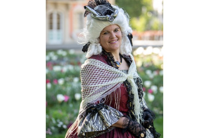 Eine Frau in barockem Kost&uuml;m mit wei&szlig;er Per&uuml;cke und rotem Kleid steht l&auml;chelnd im Freien vor einem Blumenbeet., &copy; Touristik und Marketing GmbH