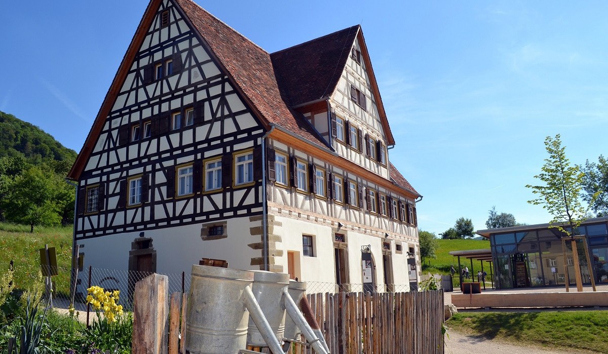 Fachwerkhaus mit spitzem Dach, umgeben von Natur. Im Vordergrund ein Holzzaun mit Gießkannen, im Hintergrund blauer Himmel und ein modernes Gebäude., © Bildarchiv Freilichtmuseum Beuren Fachwerkhaus mit spitzem Dach, umgeben von Natur. Im Vordergrund ein Holzzaun mit Gießkannen, im Hintergrund blauer Himmel und ein modernes Gebäude., © Bildarchiv Freilichtmuseum Beuren