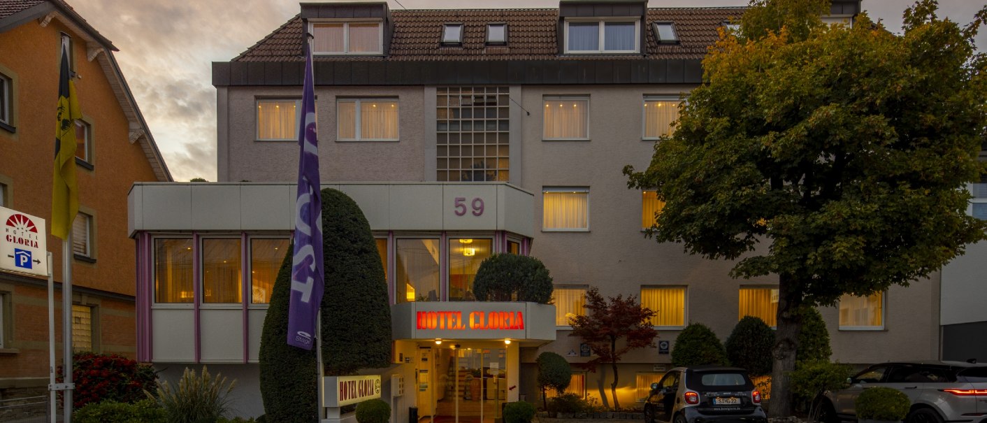 Hotelgebäude mit beleuchteten Fenstern und Eingang, umgeben von Bäumen und geparkten Autos bei Dämmerung., © TOMAS