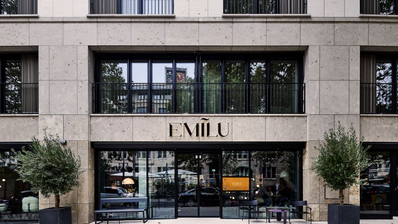 Eingang des EmiLu Hotels mit großen Glasfenstern, Pflanzen in schwarzen Töpfen und dem Hotelnamen über der Tür., © EmiLu Hotel GmbH