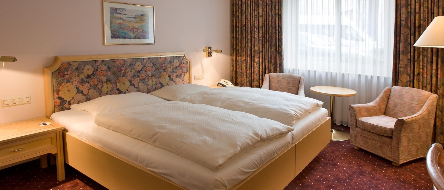 Komfortables Hotelzimmer mit Doppelbett, floralem Kopfteil, zwei Sesseln, kleinem Tisch und Fenster mit Vorhängen., © Erikson Hotel Komfortables Hotelzimmer mit Doppelbett, floralem Kopfteil, zwei Sesseln, kleinem Tisch und Fenster mit Vorhängen., © Erikson Hotel