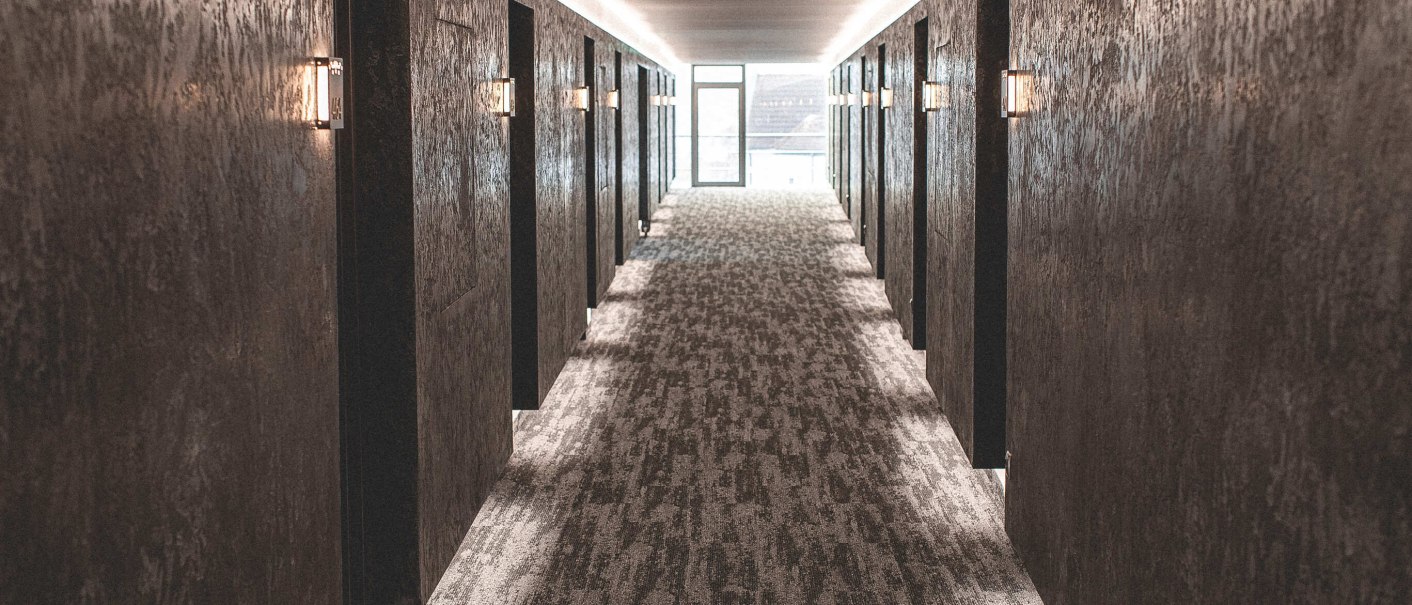 Langer, moderner Flur mit dunklen Wänden und Teppichboden, beleuchtet durch Wandlampen, führt zu einer Glastür am Ende., © Plaza Residence Stuttgart-Ditzingen Langer, moderner Flur mit dunklen Wänden und Teppichboden, beleuchtet durch Wandlampen, führt zu einer Glastür am Ende., © Plaza Residence Stuttgart-Ditzingen