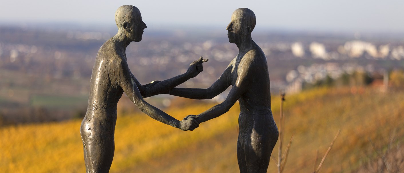 Zwei Bronzeskulpturen halten sich die Hände auf einem Hügel. Der Hintergrund ist unscharf und zeigt eine Landschaft., © SMG, Achim Mende Zwei Bronzeskulpturen halten sich die Hände auf einem Hügel. Der Hintergrund ist unscharf und zeigt eine Landschaft., © SMG, Achim Mende