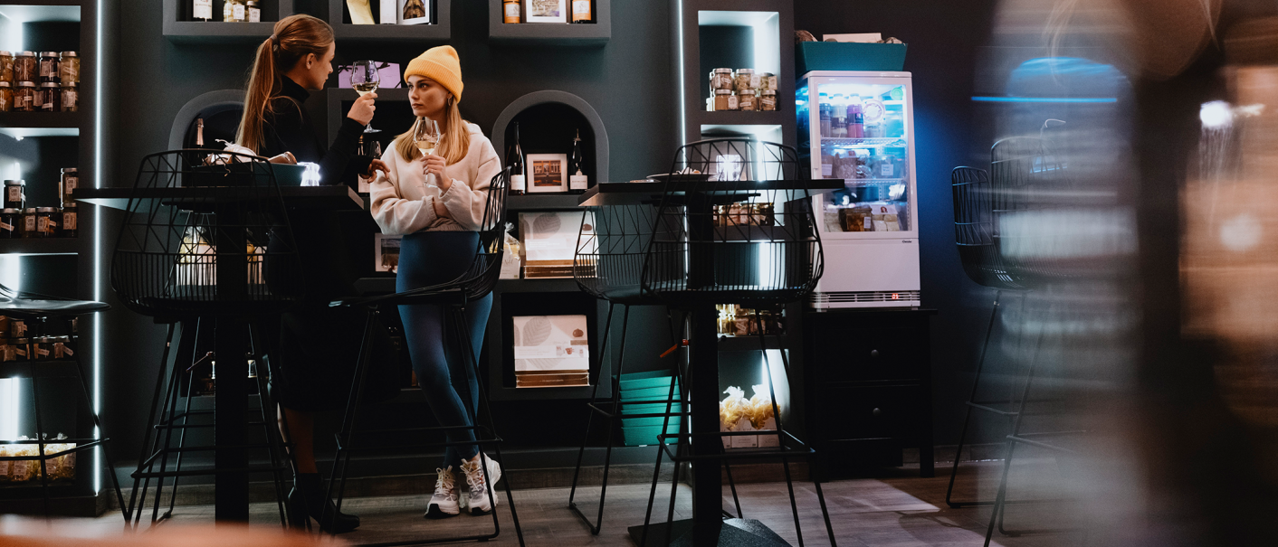 Zwei Frauen in einem stilvollen Café, eine mit gelber Mütze, halten Weingläser. Im Hintergrund sind Regale mit Produkten und ein Kühlschrank zu sehen., © Stuttgart-Marketing GmbH, Alwin Maigler Zwei Frauen in einem stilvollen Café, eine mit gelber Mütze, halten Weingläser. Im Hintergrund sind Regale mit Produkten und ein Kühlschrank zu sehen., © Stuttgart-Marketing GmbH, Alwin Maigler