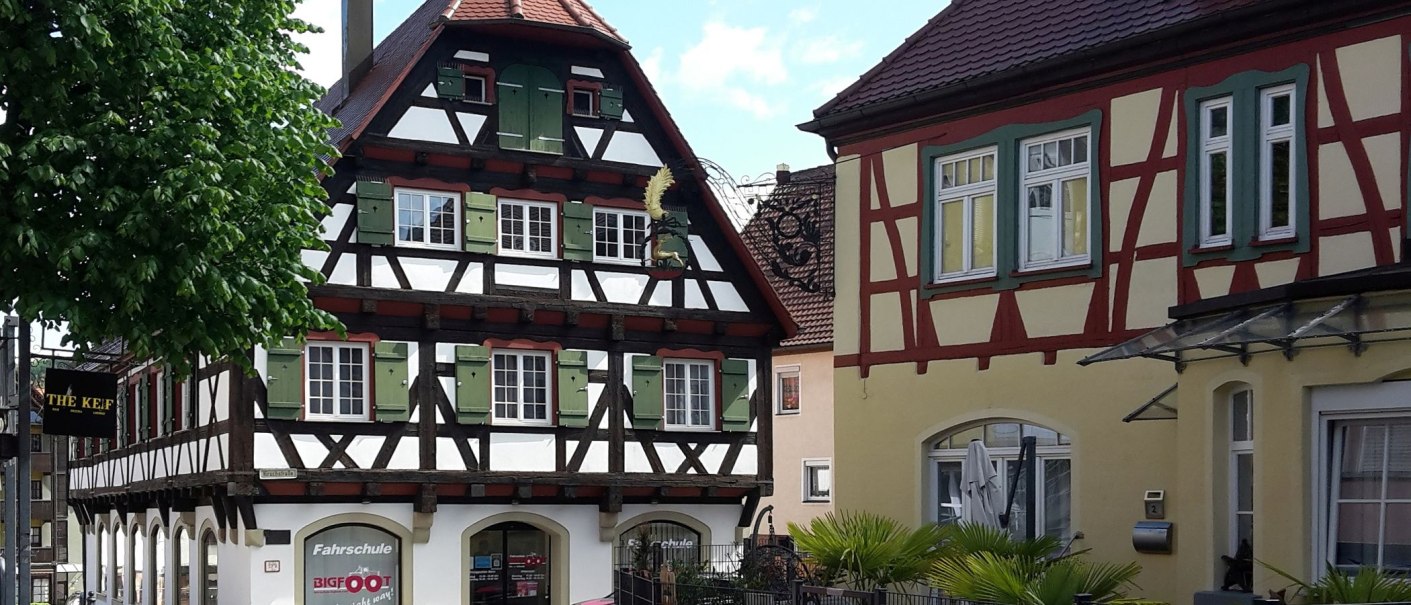 Alter Speicher - Fachwerkhaus mit dunklem Holz, weißen Gefachen und grünen Fensterläden in der Bildmitte, rechts daneben ein gelbes Haus mit rot-braunem Fachwerk im Obergeschoß und altem Wirtshausschild, © Petra Natzkowski