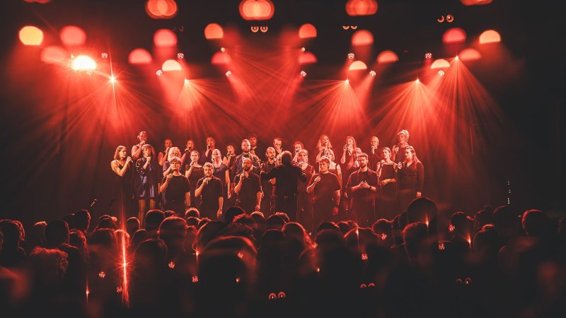 Ein A-Cappella-Chor performt auf einer B&uuml;hne mit intensiver roter Beleuchtung. Die S&auml;nger stehen in mehreren Reihen und singen vor einem gro&szlig;en Publikum.