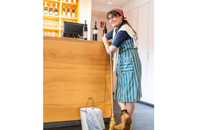 Frau in traditioneller Kleidung lehnt lächelnd an einer Theke mit Weinflaschen. Ein Eimer und Besen stehen daneben., © Stuttgart Marketing GmbH Frau in traditioneller Kleidung lehnt lächelnd an einer Theke mit Weinflaschen. Ein Eimer und Besen stehen daneben., © Stuttgart Marketing GmbH