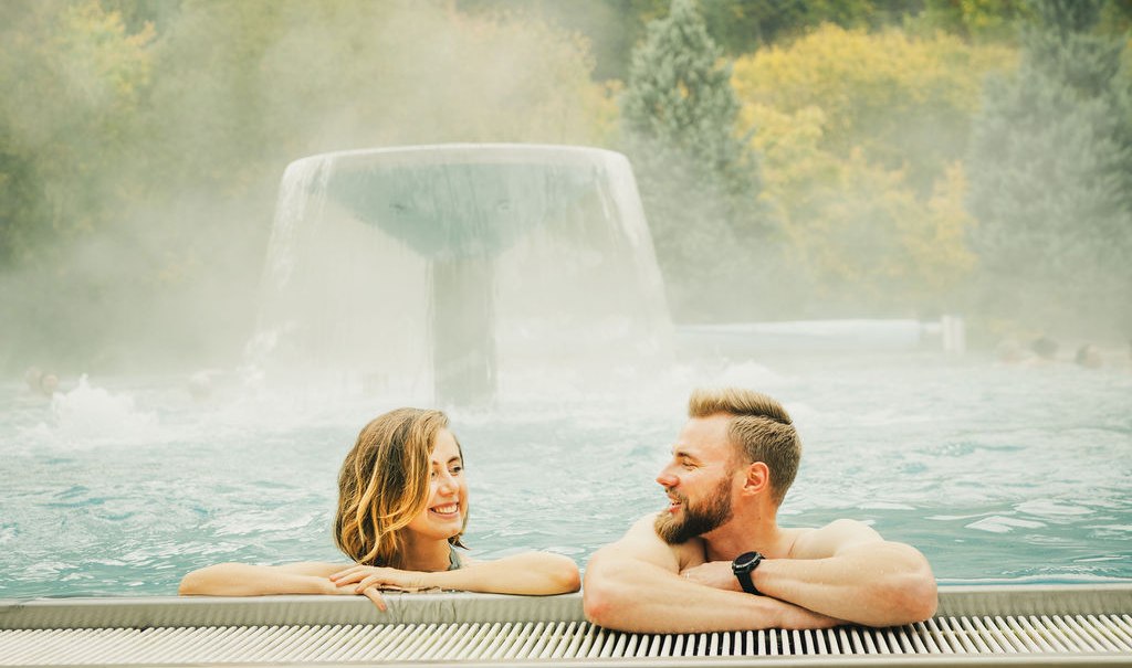 Zwei Personen entspannen in einem Au&szlig;enpool der Paracelsus-Therme Bad Liebenzell. Im Hintergrund ein Wasserfall und bewaldete H&uuml;gel., &copy; LOCHER Fotodesign & Manufaktur