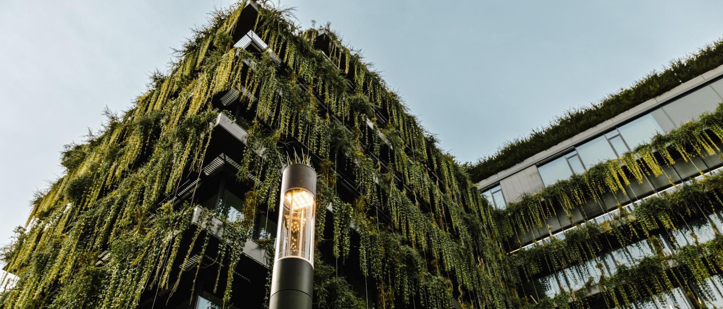 Ein modernes Gebäude mit einer grünen Fassade, bedeckt von hängenden Pflanzen. Im Vordergrund steht eine beleuchtete Straßenlaterne., © Stuttgart-Marketing GmbH, Sarah Schmid