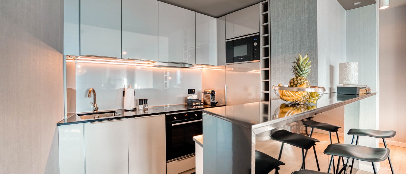 Moderne Küche mit weißen Hochglanzschränken, eingebautem Ofen und Mikrowelle. Eine Theke mit Barhockern und einer Obstschale mit Ananas., © CLOUD N°7 APARTMENTS GmbH
