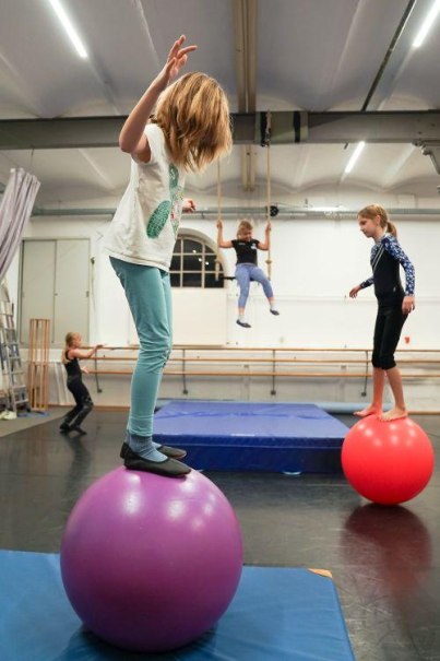 Kinder balancieren auf bunten Gymnastikbällen in einem Zirkustrainingsraum. Im Hintergrund schwingt ein Kind an einem Trapez., © Kunstschule Labyrinth Kinder balancieren auf bunten Gymnastikbällen in einem Zirkustrainingsraum. Im Hintergrund schwingt ein Kind an einem Trapez., © Kunstschule Labyrinth