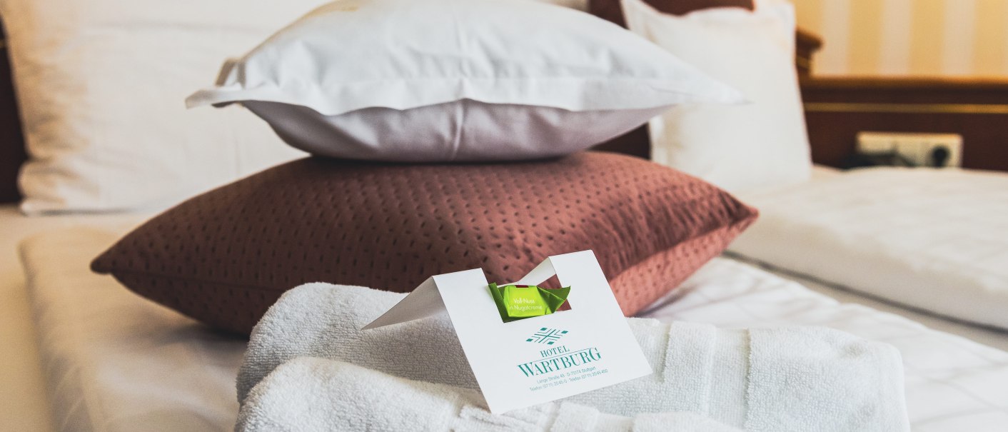 Ein Hotelbett mit mehreren Kissen und gefalteten Handtüchern. Eine Karte des Hotel Wartburg liegt auf den Handtüchern., © TOMAS