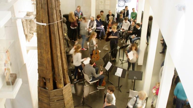 Menschen versammeln sich in einem modernen Raum mit hohen Decken. Ein gro&szlig;es Holzobjekt steht zentral. Einige Personen sitzen, andere stehen und unterhalten sich., &copy; Ludwigsburg Museum