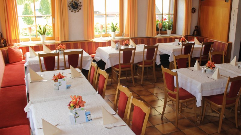 Gemütlicher Gastraum mit gedeckten Tischen, roten Stühlen und orangenen Vorhängen. Pflanzen und Dekorationen verleihen eine einladende Atmosphäre., © Gasthof zur Rose