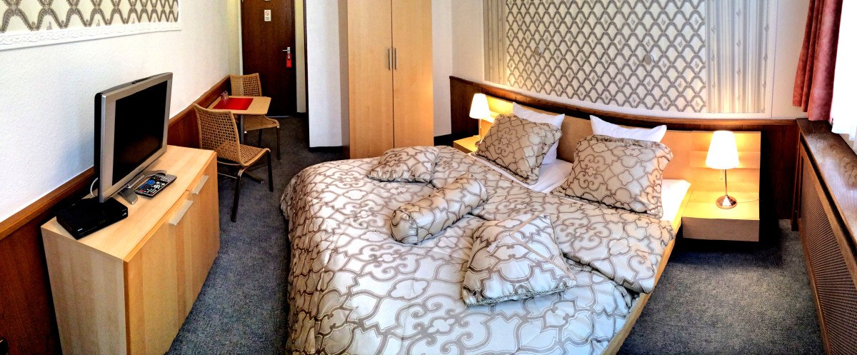 Hotelzimmer mit Doppelbett, TV, Schreibtisch und Stuhl. Gemusterte Tapete und Bettwäsche, warme Beleuchtung. Hotelzimmer mit Doppelbett, TV, Schreibtisch und Stuhl. Gemusterte Tapete und Bettwäsche, warme Beleuchtung.