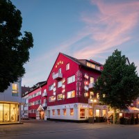 Rotes Hotelgebäude mit Flaggen und beleuchteten Schildern bei Dämmerung, umgeben von Bäumen und Straßenlaternen., © Hetzel Hotel Stuttgart