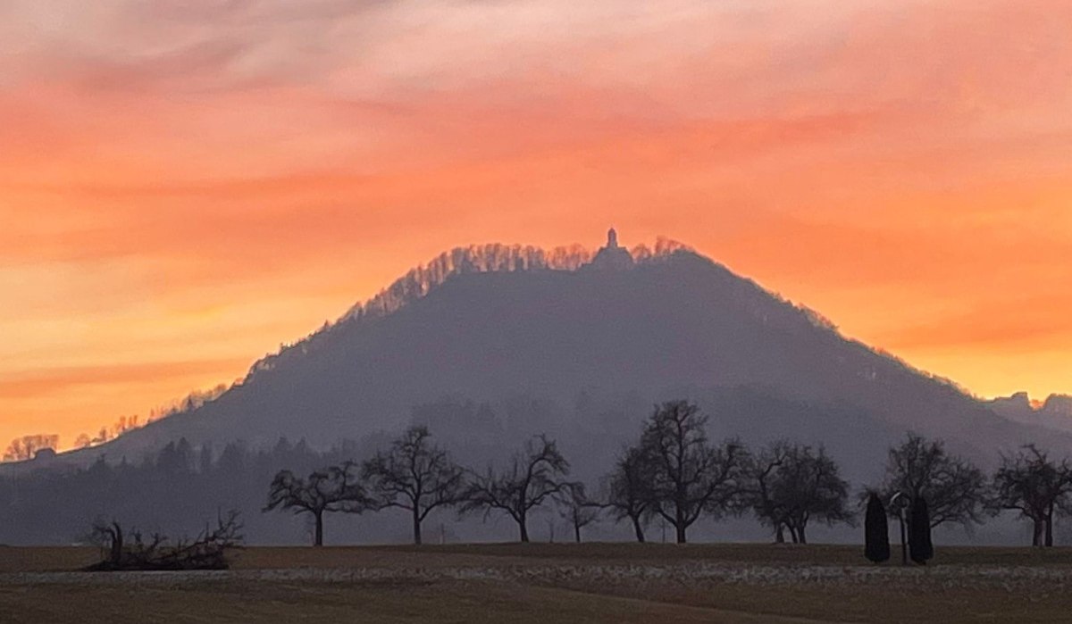 Hügel im Sonnenuntergang mit Silhouette einer Struktur auf der Spitze, umgeben von kahlen Bäumen und einem orangefarbenen Himmel., © Foto: Cornelia Steinbach Hügel im Sonnenuntergang mit Silhouette einer Struktur auf der Spitze, umgeben von kahlen Bäumen und einem orangefarbenen Himmel., © Foto: Cornelia Steinbach