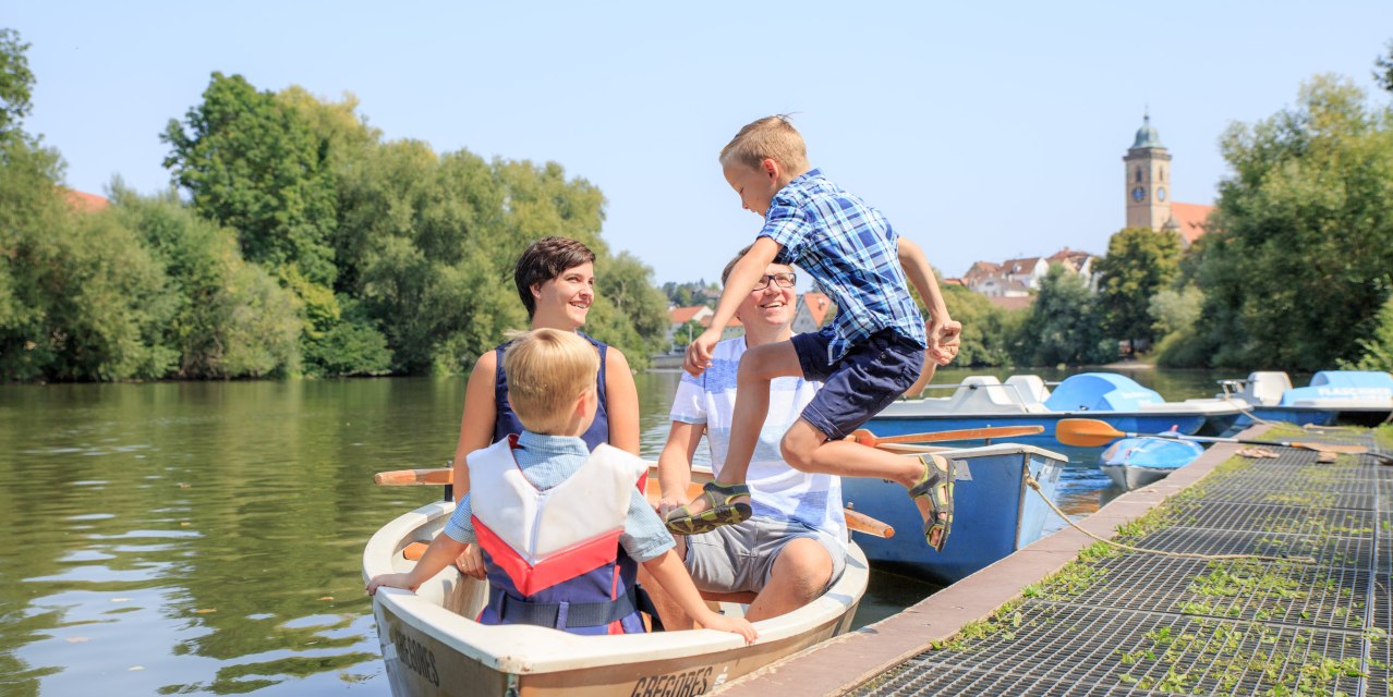 Eine Familie sitzt in einem Ruderboot auf dem Neckar. Ein Junge springt ins Boot. Im Hintergrund sind Bäume und ein Kirchturm zu sehen., © H. Bergmüller Eine Familie sitzt in einem Ruderboot auf dem Neckar. Ein Junge springt ins Boot. Im Hintergrund sind Bäume und ein Kirchturm zu sehen., © H. Bergmüller