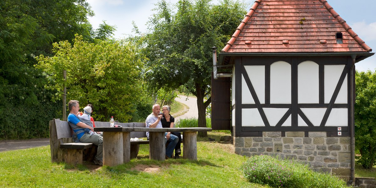 Drei Personen sitzen an einem Holztisch neben einem kleinen Fachwerkhaus im Gr&uuml;nen. Es ist ein sonniger Tag., &copy; Corinna Jacobs