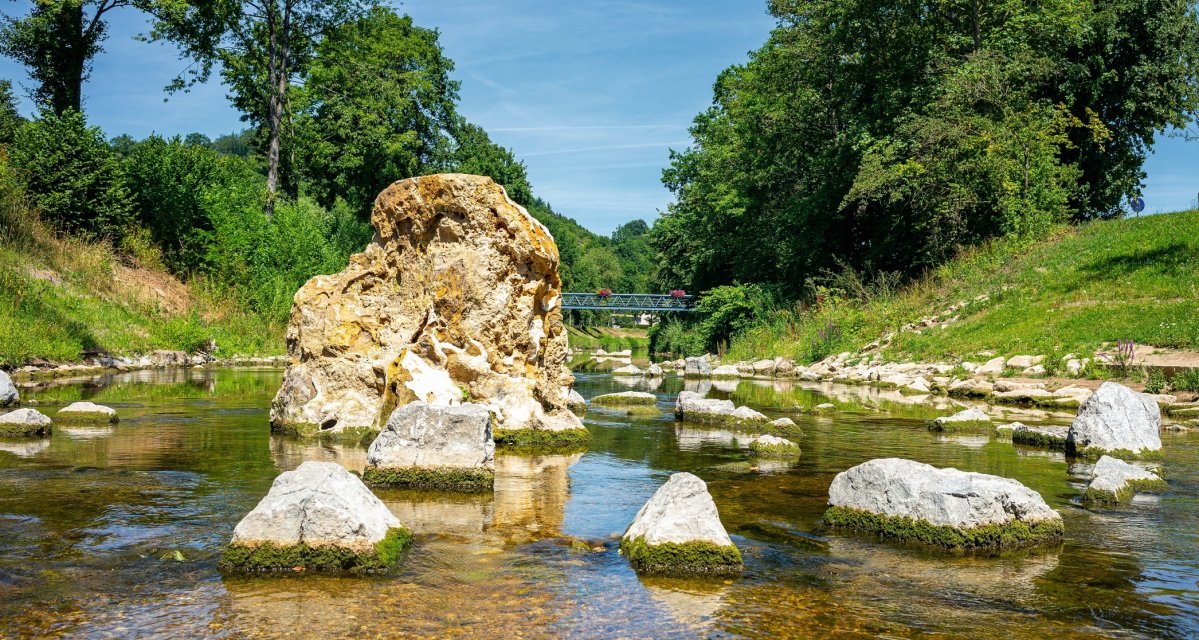 Ein Fluss mit gro&szlig;en Steinen im Wasser, umgeben von gr&uuml;ner Vegetation. Im Hintergrund ist eine Br&uuml;cke zu sehen., &copy; Remstal Tourismus e.V.