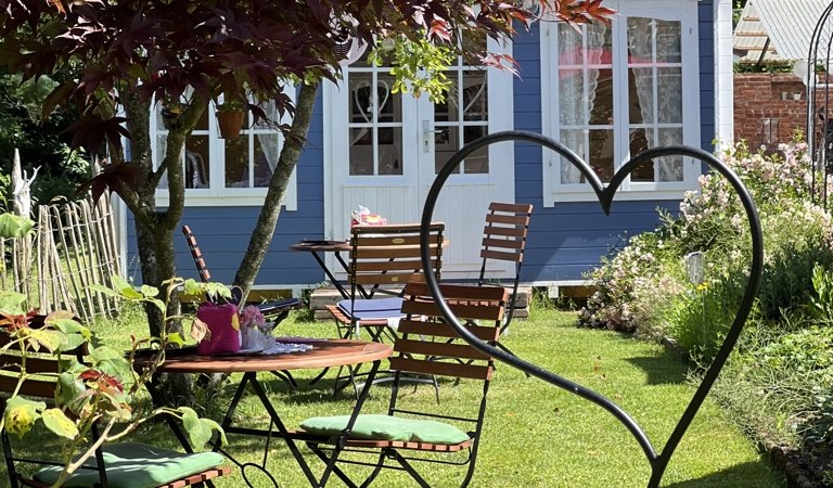 Garten mit Holztischen und -stühlen, umgeben von Pflanzen. Im Hintergrund ein blaues Häuschen und ein Baum mit roten Blättern., © Café Plätschwiesle