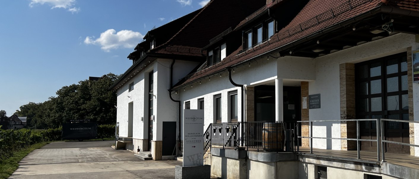 Weißes Gebäude mit rotem Dach, Vinothek-Schild und Weinfass vor dem Eingang. Umgeben von Bäumen und klarem Himmel., © Weinfactum Bad Cannstatt Weißes Gebäude mit rotem Dach, Vinothek-Schild und Weinfass vor dem Eingang. Umgeben von Bäumen und klarem Himmel., © Weinfactum Bad Cannstatt