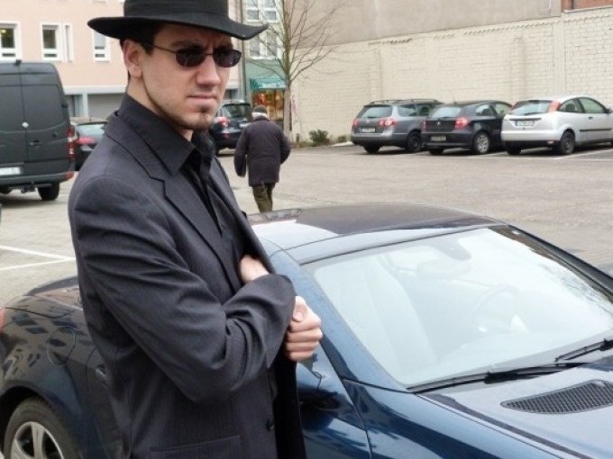 Ein Mann in dunklem Anzug und Hut steht neben einem blauen Auto auf einem Parkplatz. Im Hintergrund sind weitere Autos und Gebäude zu sehen., © Cool-Tours StattReisen Ein Mann in dunklem Anzug und Hut steht neben einem blauen Auto auf einem Parkplatz. Im Hintergrund sind weitere Autos und Gebäude zu sehen., © Cool-Tours StattReisen