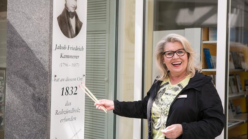 Eine Frau steht lächelnd vor einer Tafel, die Jakob Friedrich Kammerer und seine Erfindung des Reibzündholzes 1832 würdigt., © © T&E, Benjamin Stollenberg Eine Frau steht lächelnd vor einer Tafel, die Jakob Friedrich Kammerer und seine Erfindung des Reibzündholzes 1832 würdigt., © © T&E, Benjamin Stollenberg