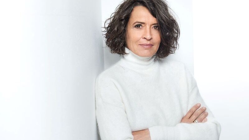 Eine Frau mit lockigem Haar in einem weißen Rollkragenpullover lehnt entspannt an einer weißen Wand und schaut in die Kamera., © Kulturbüro Schwäbisch Gmünd