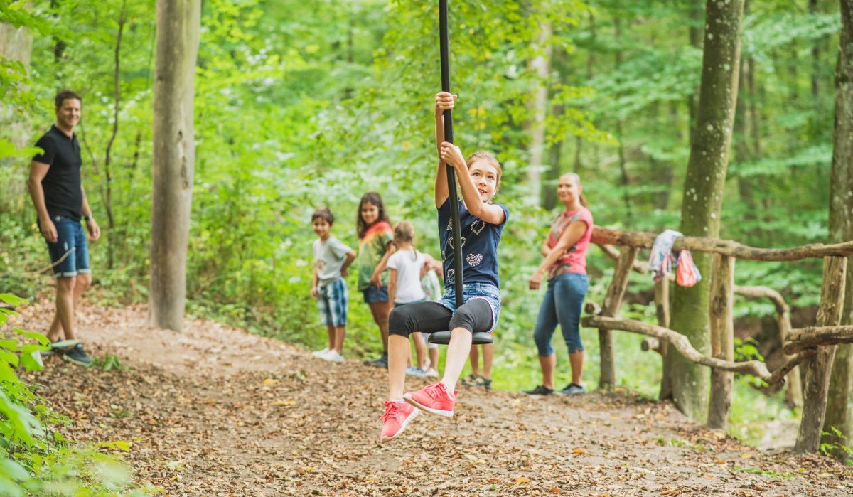 Ein Kind schwingt an einer Seilrutsche im Wald, umgeben von anderen Kindern und Erwachsenen, die zuschauen. Der Spielplatz liegt im Grünen. Ein Kind schwingt an einer Seilrutsche im Wald, umgeben von anderen Kindern und Erwachsenen, die zuschauen. Der Spielplatz liegt im Grünen.