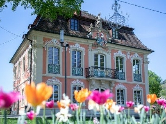 Ein barockes Gebäude mit verzierten Fenstern und Balkon, umgeben von bunten Tulpen im Vordergrund. Ein Kronleuchter hängt im Freien., © Touristik und Marketing GmbH Ein barockes Gebäude mit verzierten Fenstern und Balkon, umgeben von bunten Tulpen im Vordergrund. Ein Kronleuchter hängt im Freien., © Touristik und Marketing GmbH