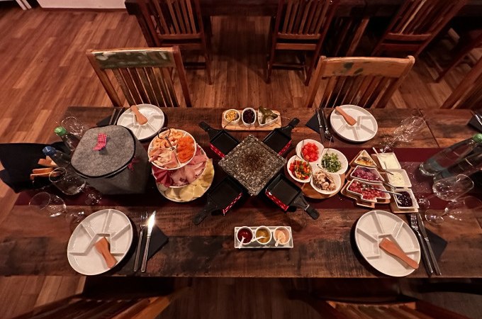 Ein Tisch mit Raclette-Grill, Tellern, Besteck und verschiedenen Zutaten für einen Raclette-Abend, darunter Gemüse, Fleisch und Saucen., © Cool-Tours StattReisen Ein Tisch mit Raclette-Grill, Tellern, Besteck und verschiedenen Zutaten für einen Raclette-Abend, darunter Gemüse, Fleisch und Saucen., © Cool-Tours StattReisen