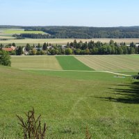Grüne Wiesen und Felder erstrecken sich bis zu einem Dorf am Waldrand unter blauem Himmel., © Foto: Cornelia Steinbach Grüne Wiesen und Felder erstrecken sich bis zu einem Dorf am Waldrand unter blauem Himmel., © Foto: Cornelia Steinbach