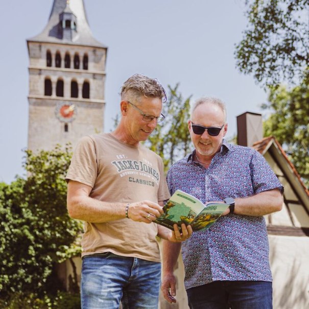 Zwei M&auml;nner stehen im Freien und schauen in ein Buch. Im Hintergrund ist ein Kirchturm zu sehen. Die Szene wirkt entspannt und fr&uuml;hlingshaft., &copy; Wirtschaftsf&ouml;rderung Sindelfingen GmbH