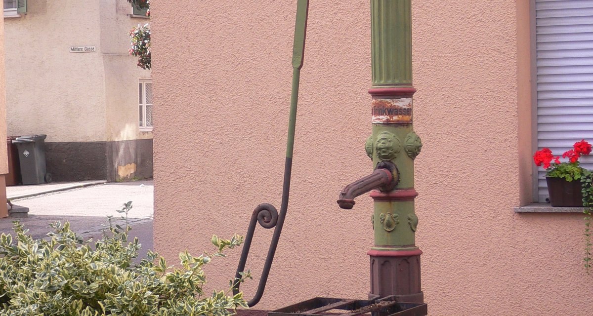 Ein grüner, dekorativer Pump-Brunnen steht vor einer rosa Hauswand. Im Vordergrund sind Pflanzen zu sehen., © Land der 1000 Hügel - Kraichgau-Stromberg Ein grüner, dekorativer Pump-Brunnen steht vor einer rosa Hauswand. Im Vordergrund sind Pflanzen zu sehen., © Land der 1000 Hügel - Kraichgau-Stromberg