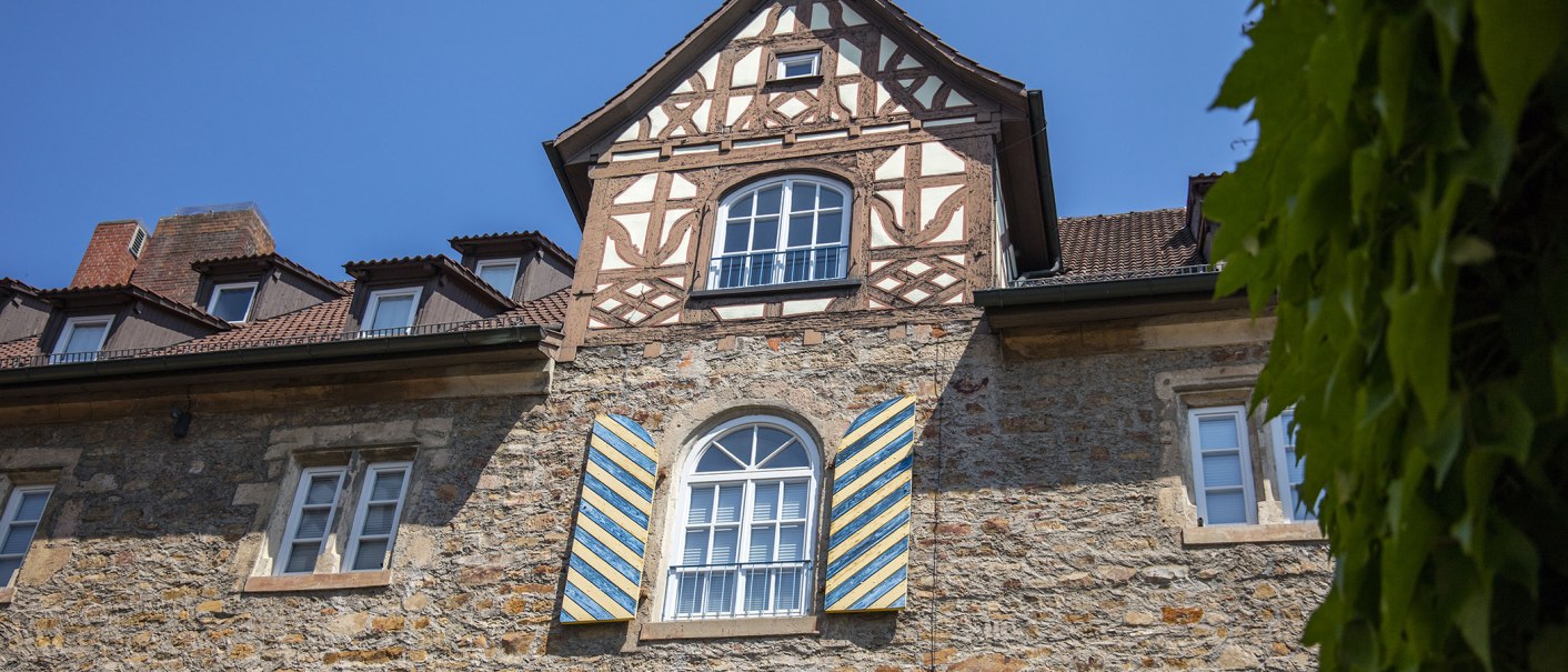 Fachwerkhaus mit blauen Fensterläden, Ziegeldach und grünem Laub im Vordergrund unter blauem Himmel., © Achim Mende Fachwerkhaus mit blauen Fensterläden, Ziegeldach und grünem Laub im Vordergrund unter blauem Himmel., © Achim Mende