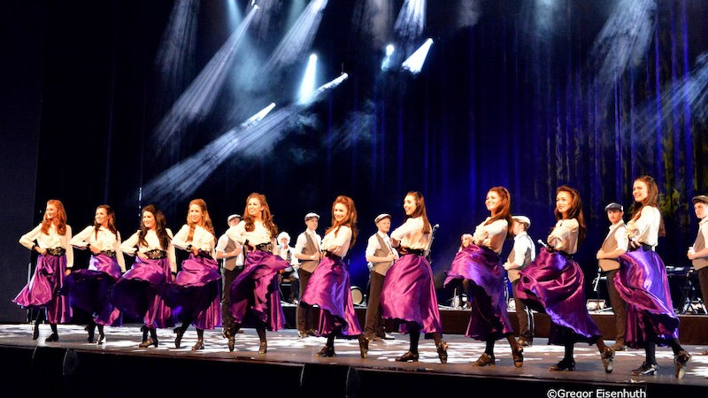Tanzgruppe in lila Röcken und weißen Blusen auf einer Bühne, beleuchtet von Scheinwerfern. Im Hintergrund sind Musiker zu sehen., © Kultur- und Sportamt Stadt Bietigheim-Bissingen
