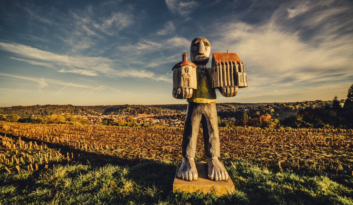 Eine Skulptur eines großen Wesens hält zwei Gebäude in den Händen, steht auf einem Feld unter einem weiten, bewölkten Himmel., © Landratsamt Ostalbkreis